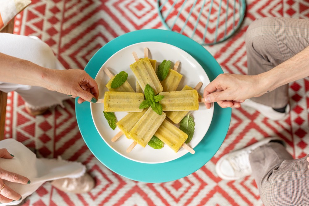 Overhead: friends reaching for house-made popsicles and mint on a teal charger, over a red patterned rug.