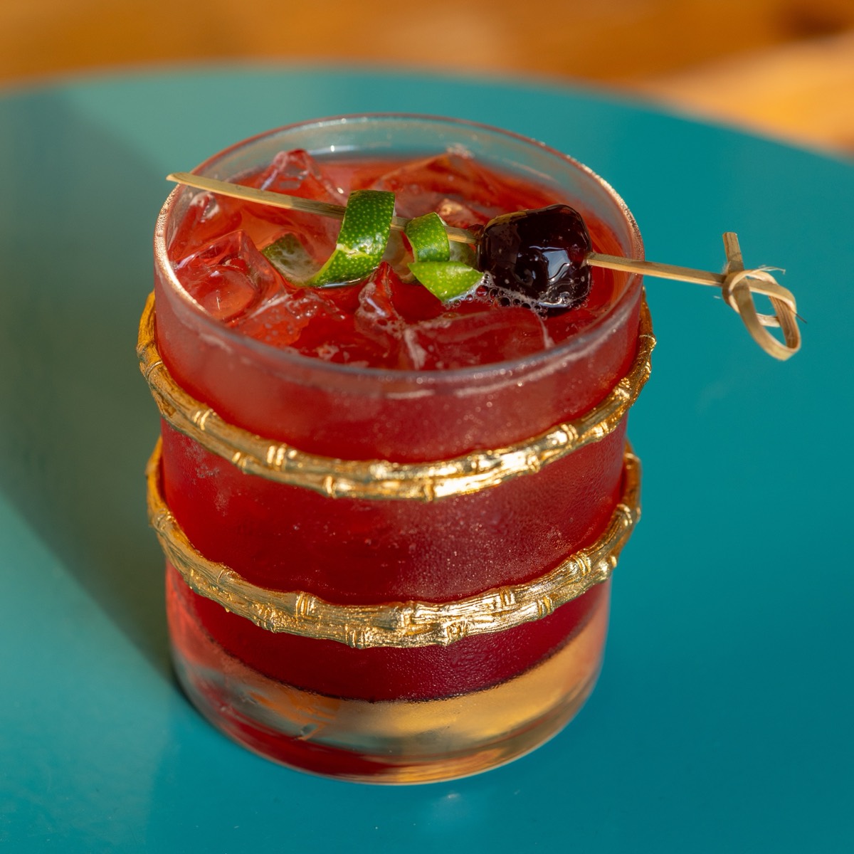 A wine-dark cocktail over ice, wrapped in gold bamboo rings, on Jar’d’s teal tabletop.